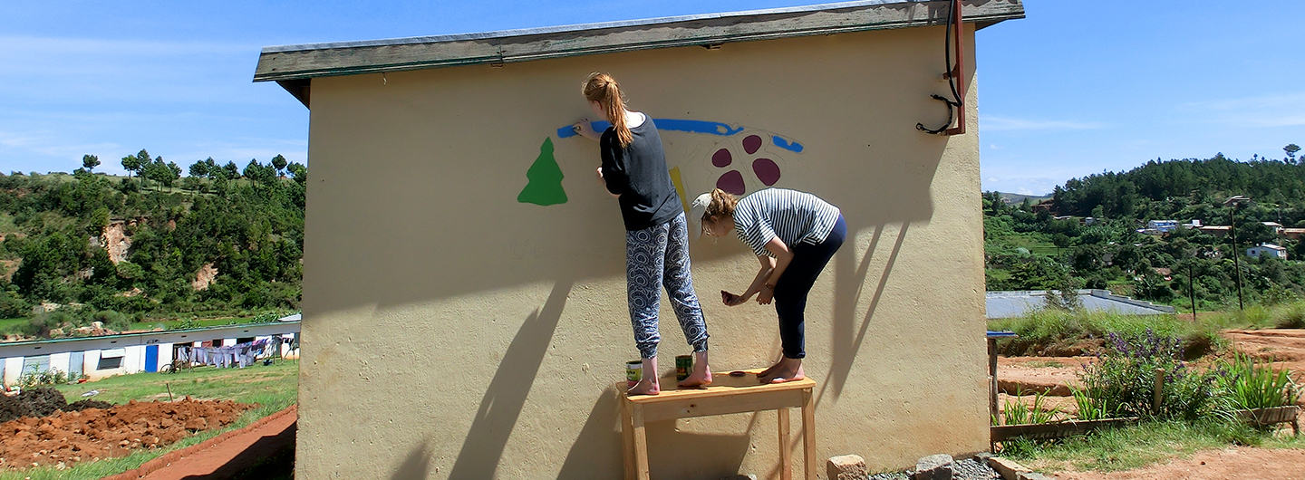 zwei junge Volontärinnen malen das Ny Hary Logo auf eine Wand in Miarinarivo in Madagaskar